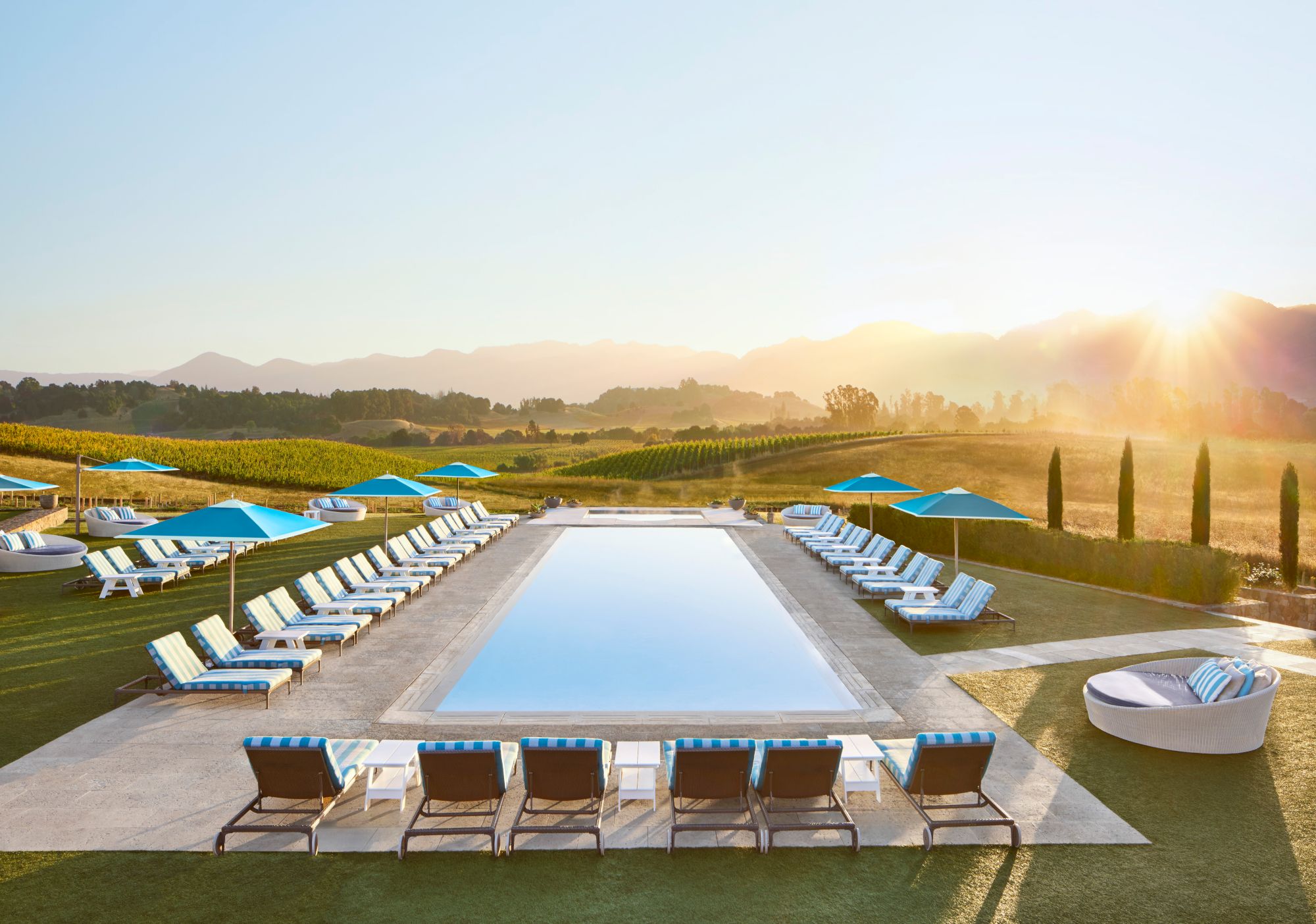 Sunlit outdoor resort: a long swimming pool, striped loungers under blue umbrellas, grassy patio, and vineyard hills at sunrise.