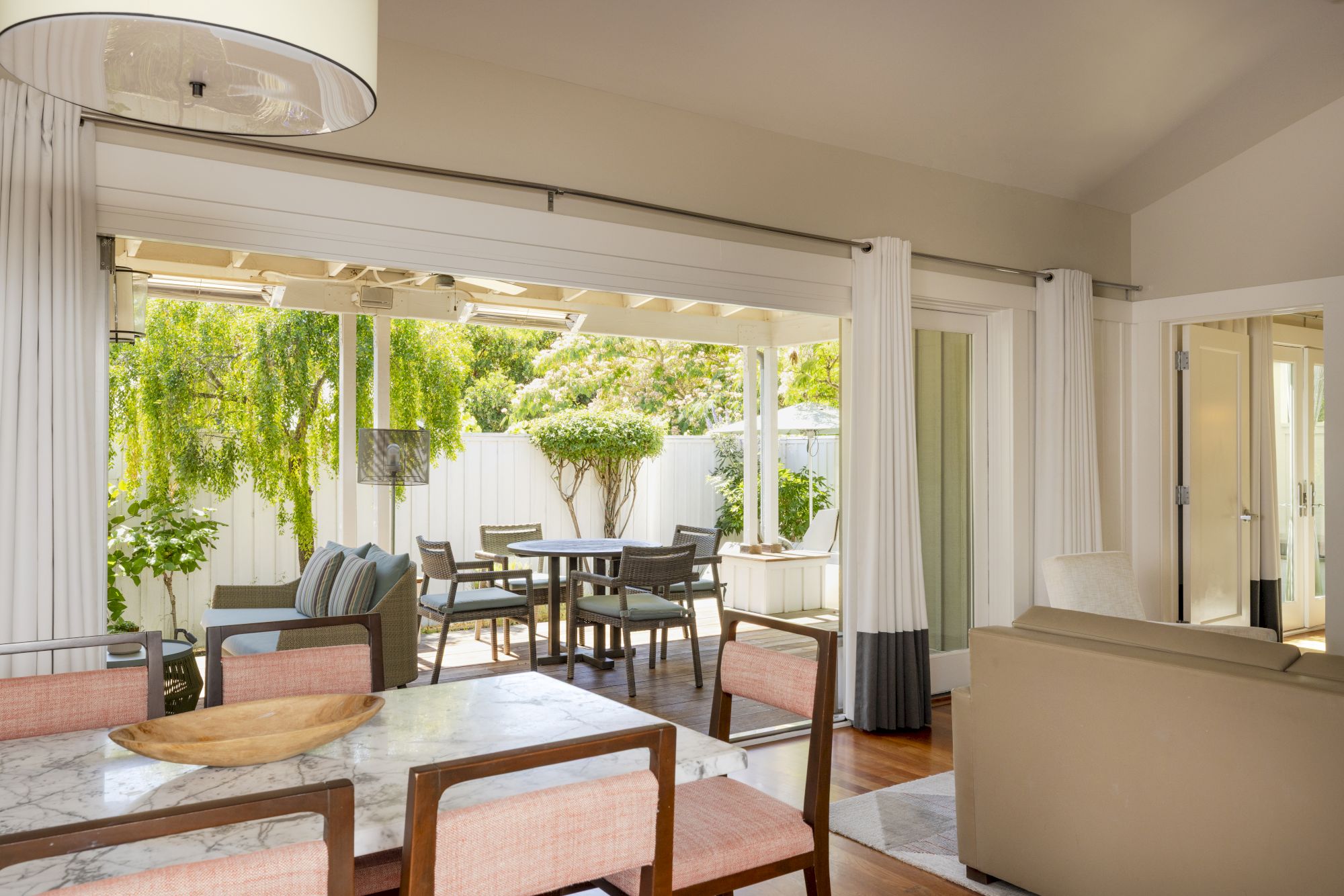 A bright living room with large sliding doors opening to a patio dining set, lush green trees, and soft curtains.