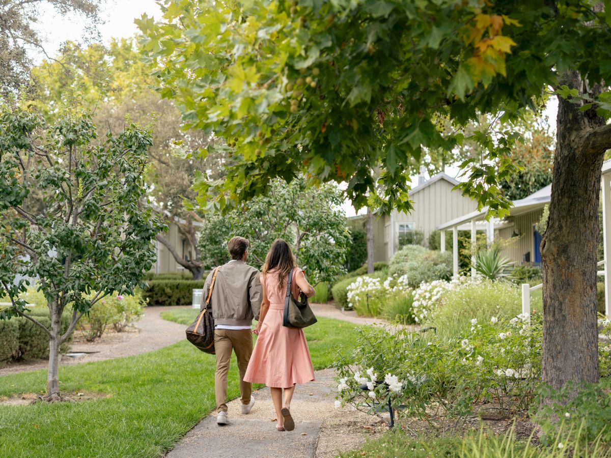 A couple strolls hand-in-hand along a sunny garden path under leafy trees near white buildings.