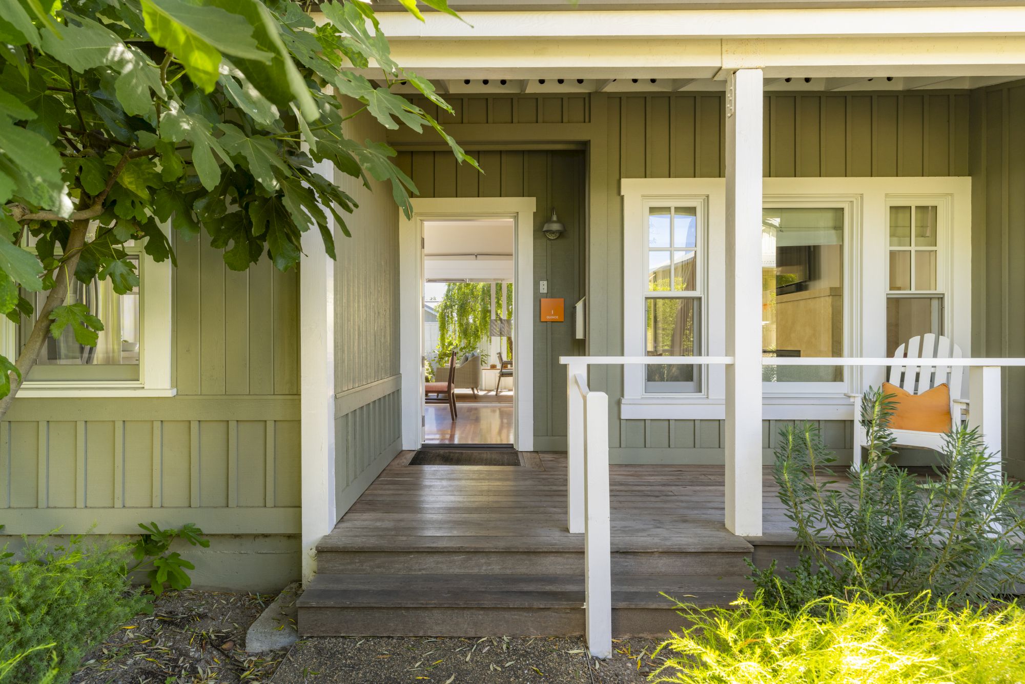 Sunlit covered porch with white columns, an open doorway, and lush green plants framing a cozy outdoor seating area.