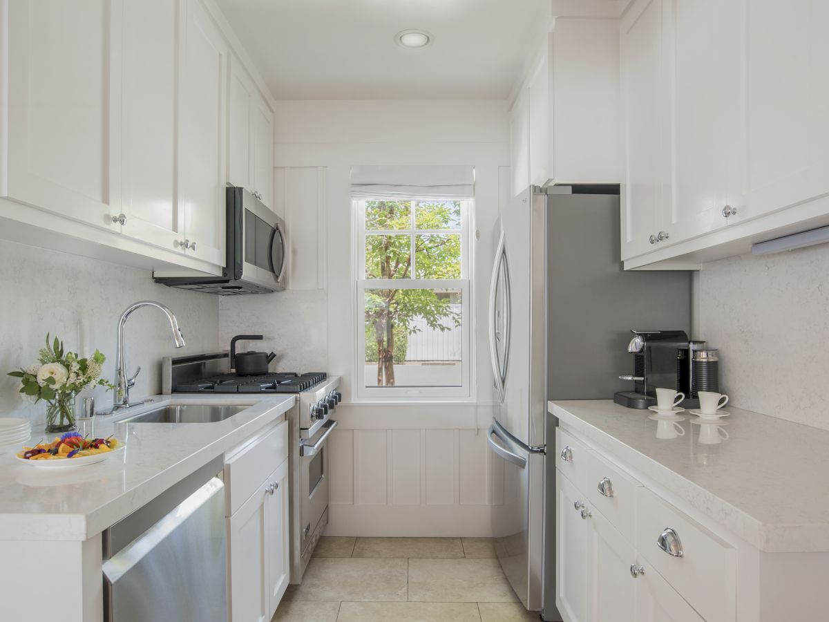 Bright modern kitchen with white cabinets, marble counters, stove, sink, window, and a coffee machine.