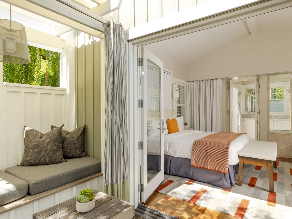 A bright modern suite shows a bed with orange throw, sheer curtains, a small seating nook, and an attached bathroom vanity with mirror.
