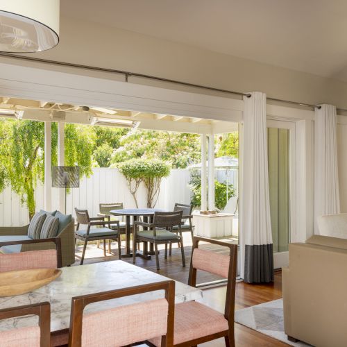 Bright patio with an outdoor table and wicker chairs, surrounded by lush green plants, visible through large sliding doors and curtains.