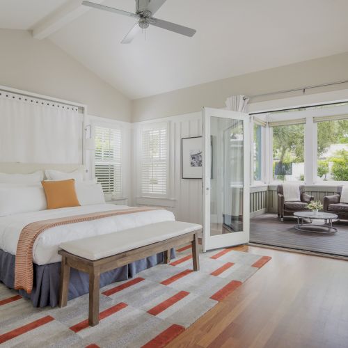 A bright bedroom with a wooden bed, ceiling fan, shuttered windows, and a sliding door leading to a seating patio area.