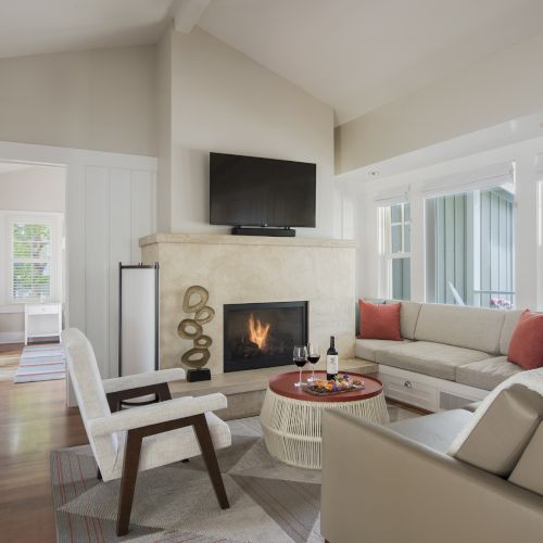 Bright modern living room with a crackling fireplace, wall-mounted TV, beige sofa, and large windows letting in daylight.