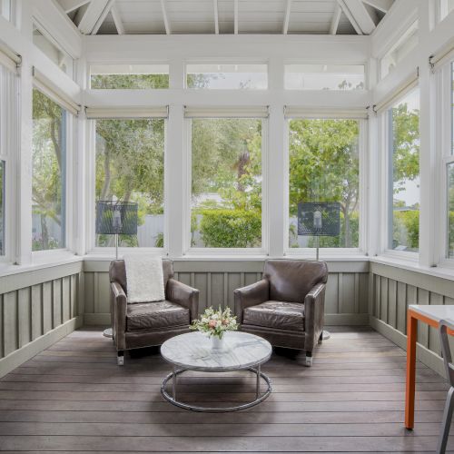 A bright sunroom with two brown leather chairs, a round marble table, and large windows overlooking lush green trees.
