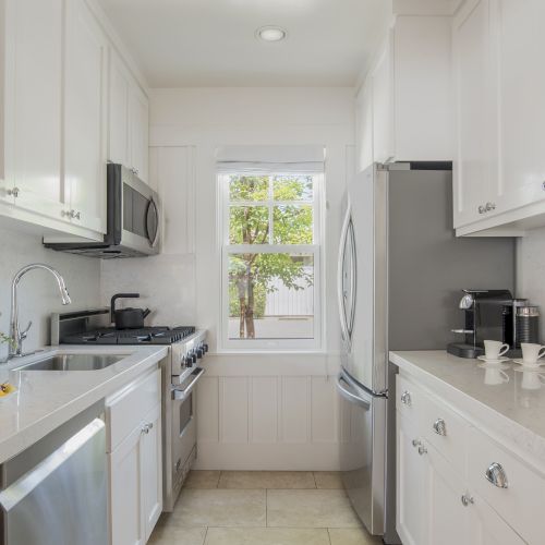 Bright modern kitchen with white cabinets, marble counters, sink, stove, window view, and a coffee machine on the counter.