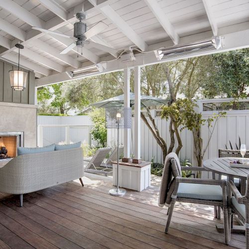 A bright covered patio with a stone fireplace, wicker chairs, and a round dining table, overlooking lush trees in the yard.