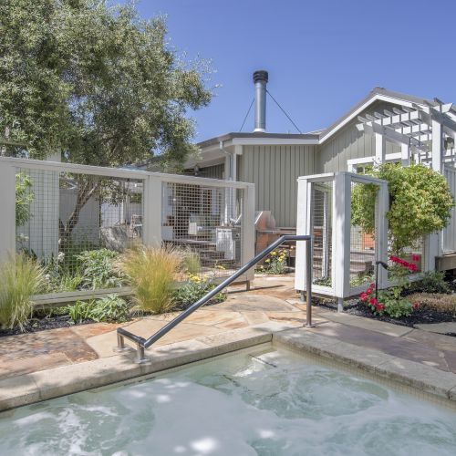 Outdoor spa area with a heated pool, stone patio, lush plants, fenced mesh, and a vent pipe under a clear blue sky.