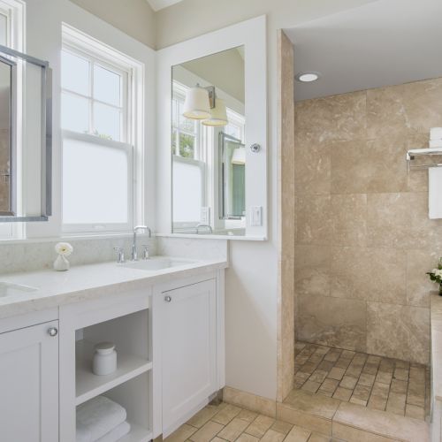 A bright modern bathroom with dual sinks, large mirrors, tiled walls, towel rack, and a glass shower enclosure.