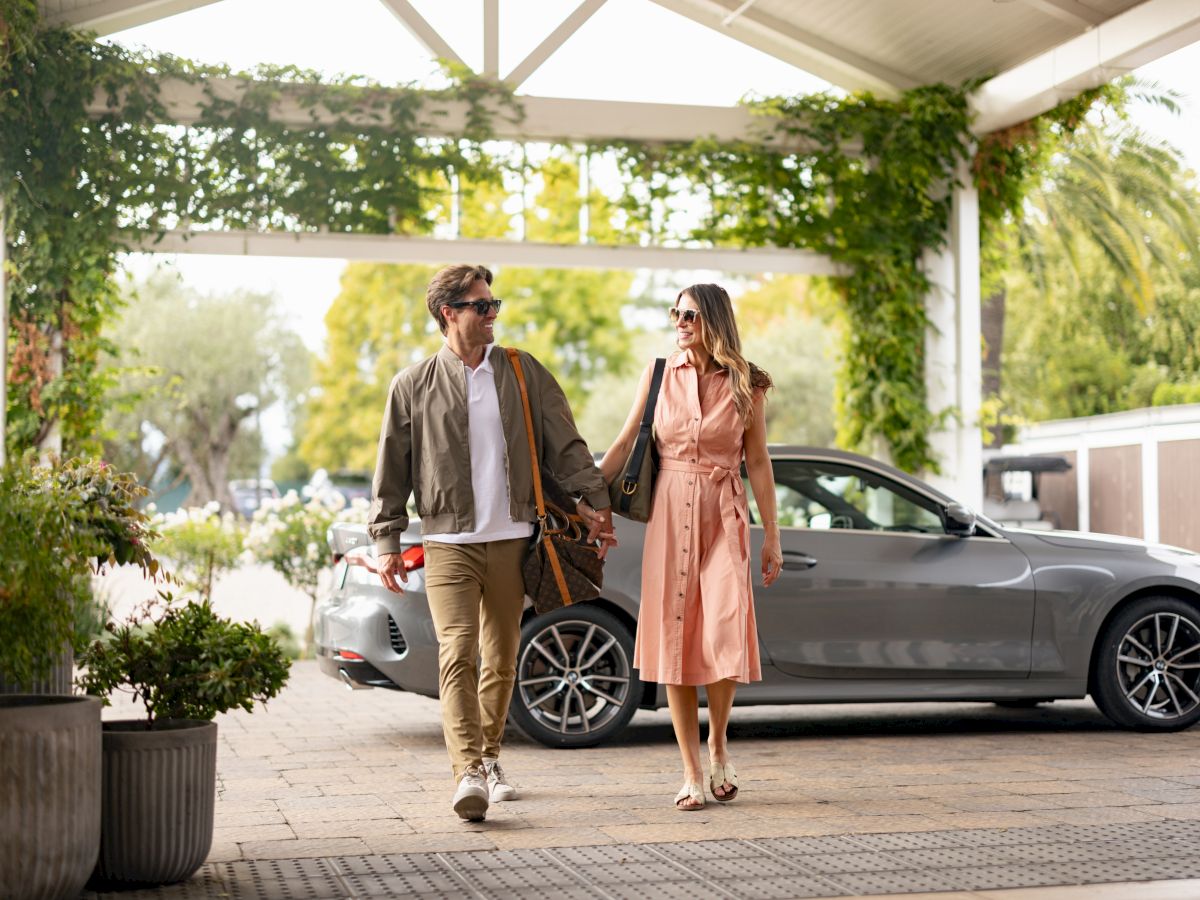 A couple strolls hand in hand beside a sleek car under a canopy, surrounded by lush greenery, smiling on vacation.