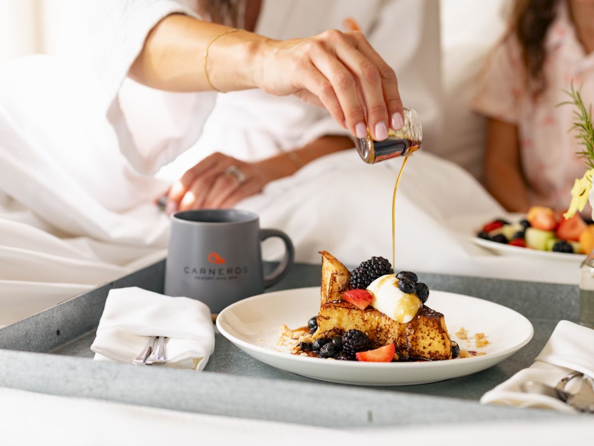 A server pours syrup over French toast topped with berries, with coffee and flowers on the table beside it.