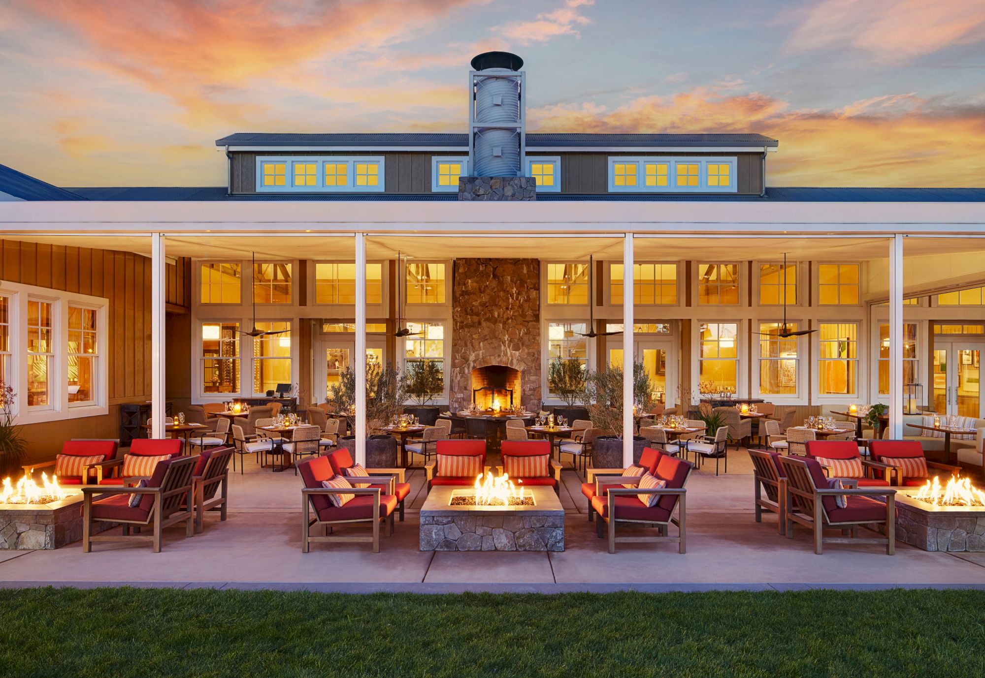 A modern hotel courtyard at sunset with outdoor lounge seating, fire pits, and warm-lit windows under colorful clouds.