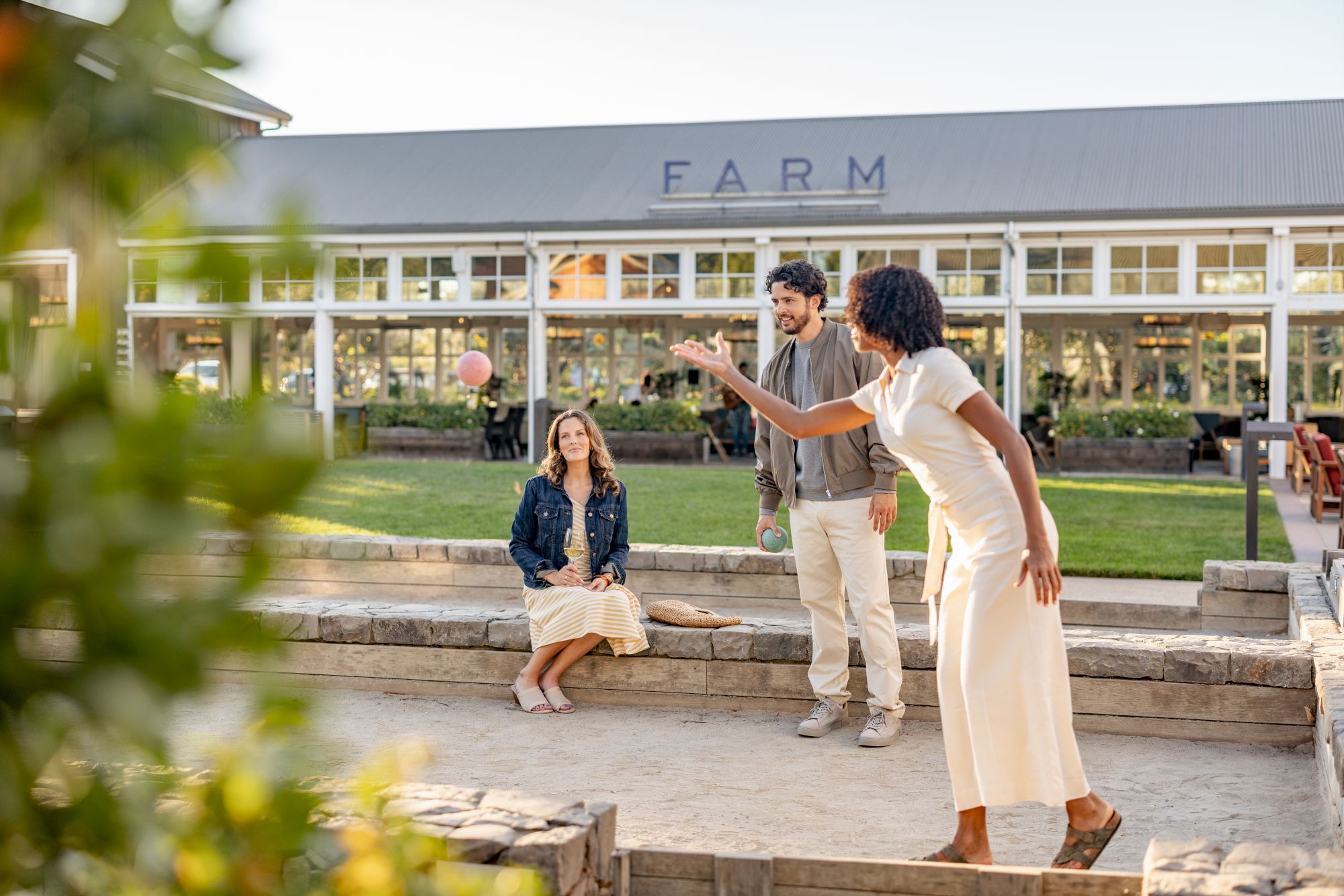 Two friends chat on a sunny patio outside a building marked &ldquo;FARM,&rdquo; with one person gesturing as others relax and smile.