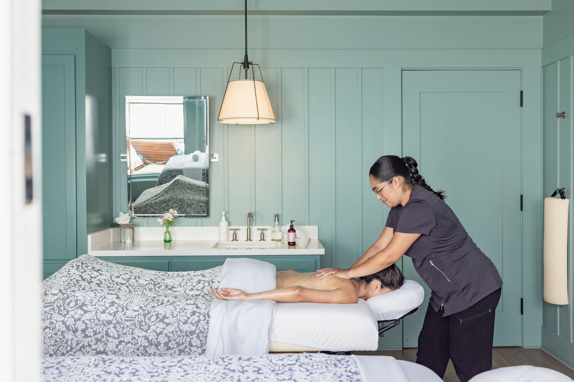 A therapist in a pastel spa room gives a relaxing massage to a reclining client on a treatment table.