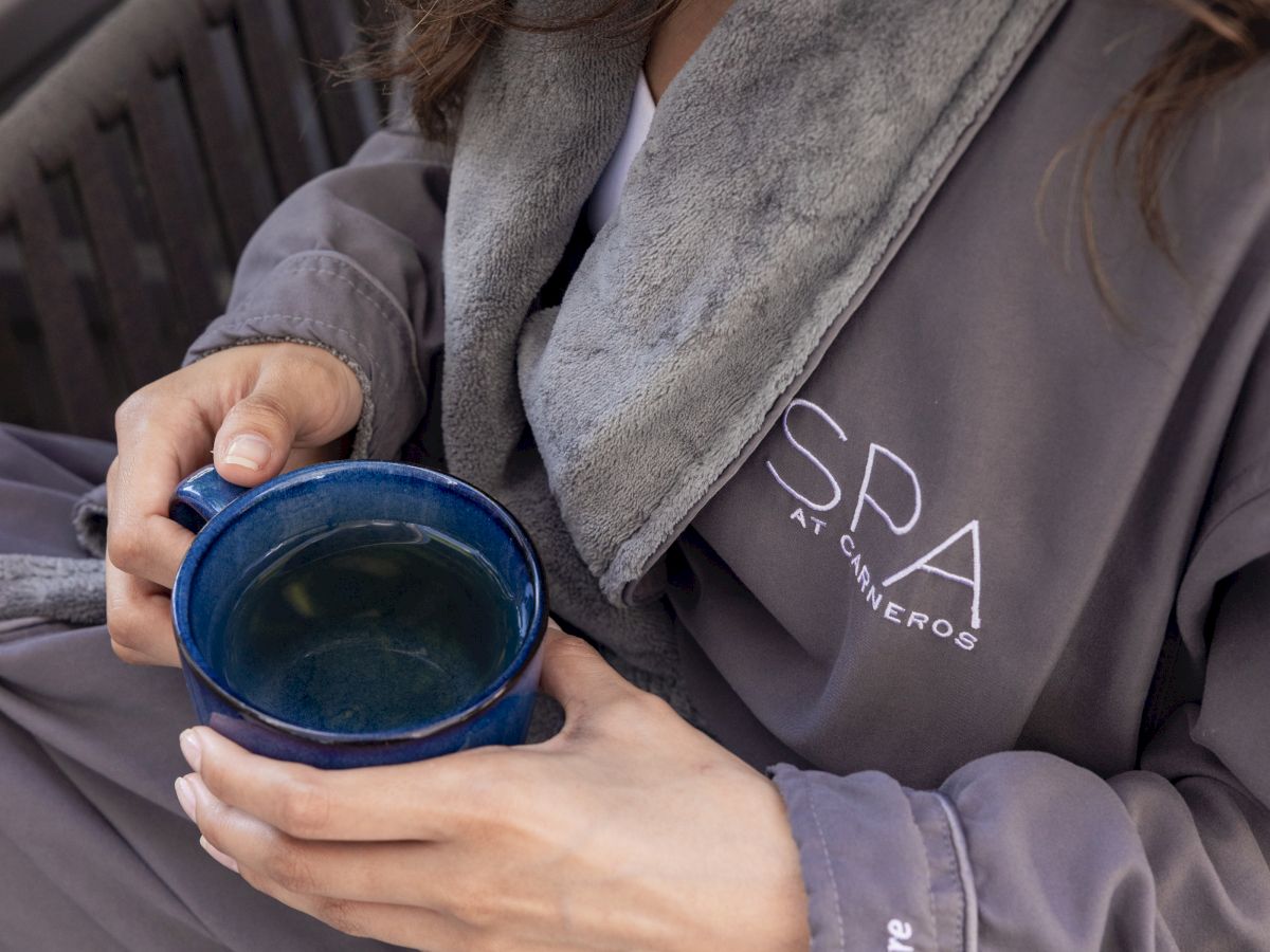 A person in a gray hoodie and robe holds a blue mug of hot drink, with cozy jewelry and warm, relaxed vibes.