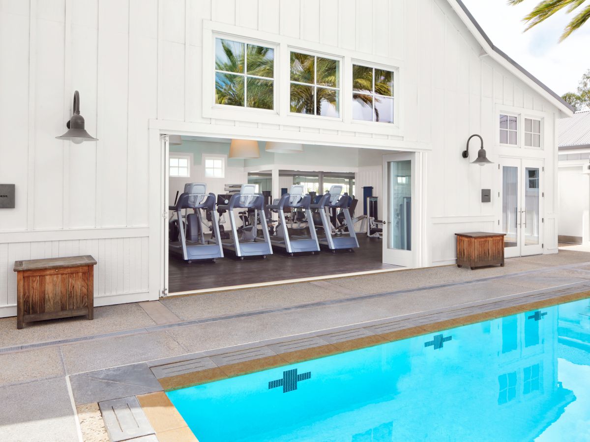 A bright hotel fitness room with treadmills and rowing machines, framed by open doors, next to a small outdoor swimming pool.