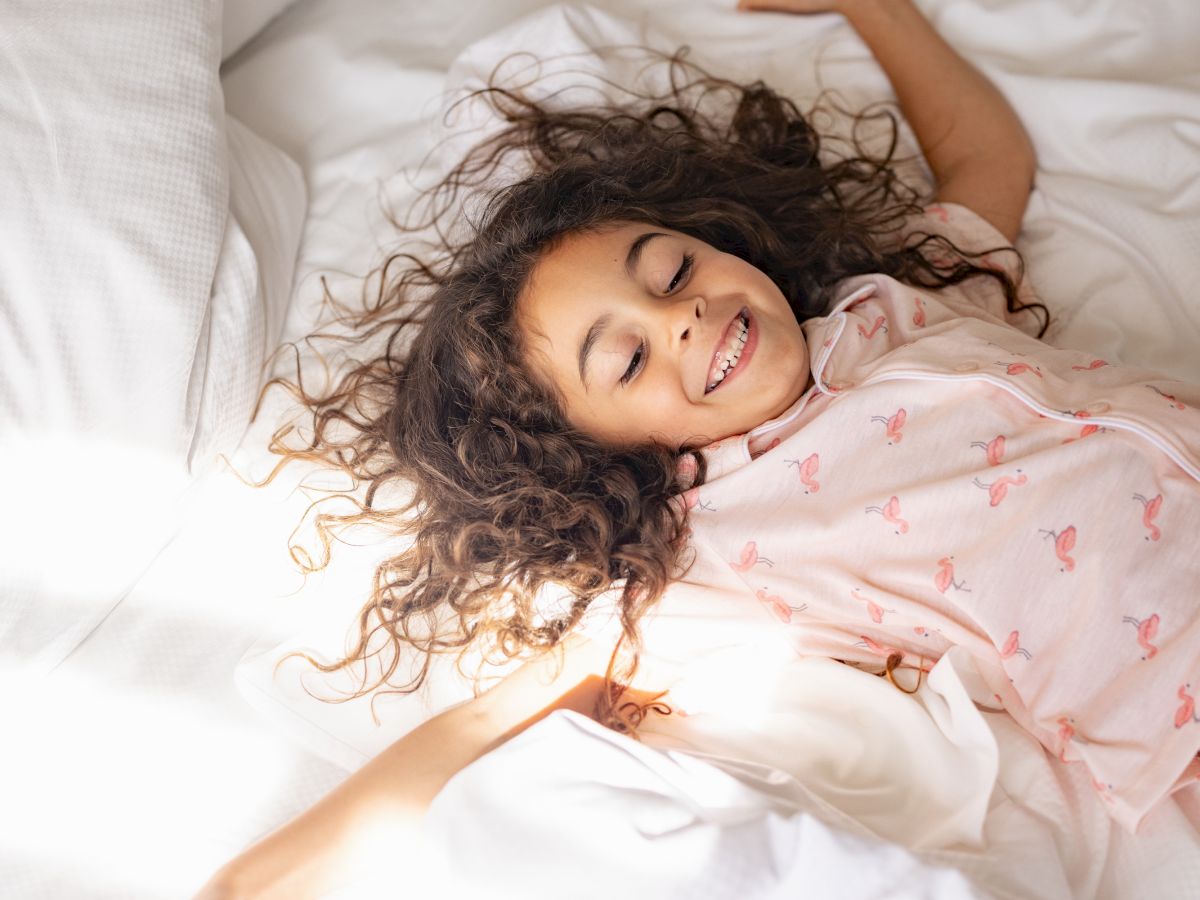 A person with curly dark hair lies on a white bed, wearing pink flamingo pajamas, smiling and slightly stretching with closed eyes.