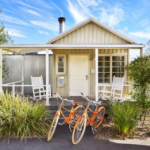 A sunny backyard patio with two white chairs, an orange bicycle, and a small modern cottage house surrounded by green plants.