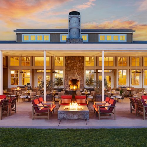 Warm sunset over a modern lodge patio with fire pits, lounge chairs, and a stone fireplace under a canopy.