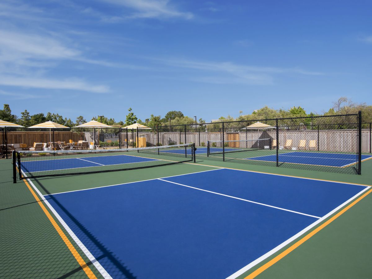 An outdoor blue pickleball/tennis court with a chain-link fence, sun umbrellas, seating, and trees under a blue sky.